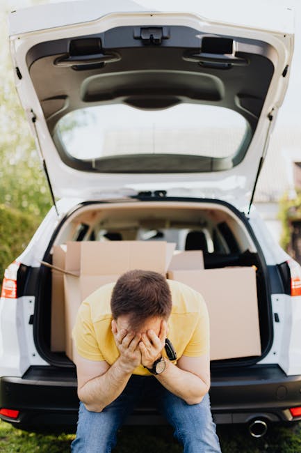A man wearing a yellow t-shirt and jeans is crouched in front of an open car trunk, covering his face with his hands. The trunk is filled with several cardboard boxes, some wrapped in clear plastic an