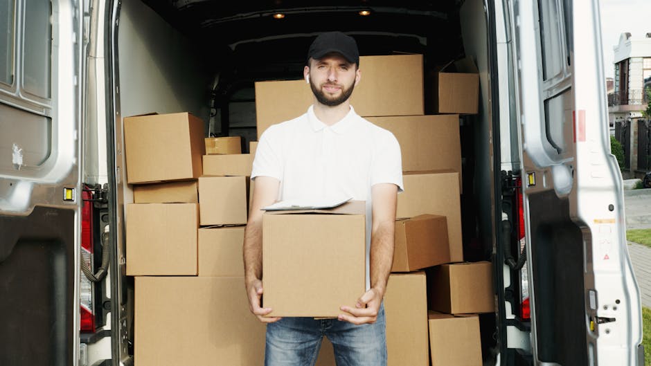 A young man with a beard wearing a black cap, white polo shirt, and jeans stands at the open back of a white moving van during daylight, holding a medium-sized cardboard box. The van is packed with se