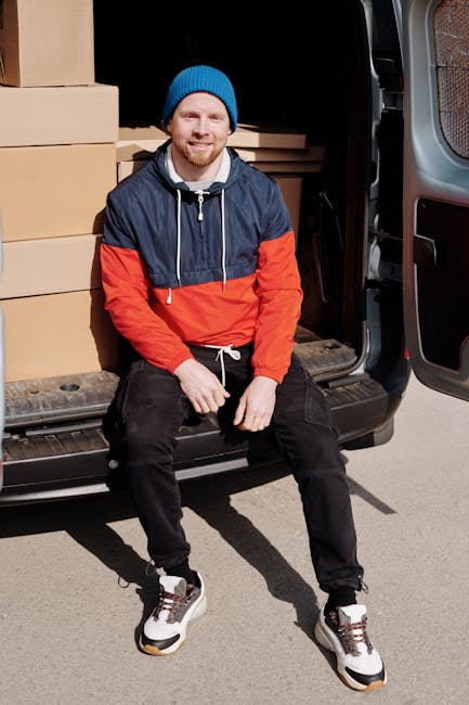 A young man with a beard wearing a black cap, white polo shirt, and jeans stands at the open back of a white moving van during daylight, holding a medium-sized cardboard box. The van is packed with se